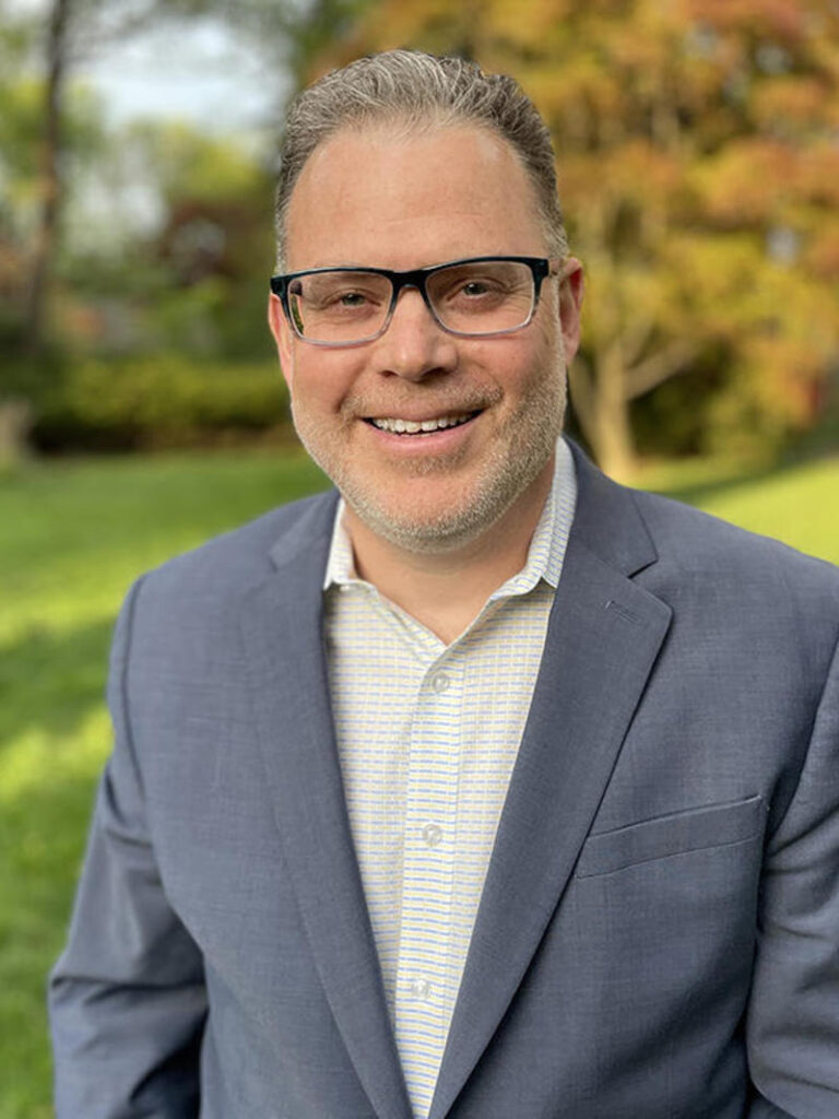 Color photograph of Bill Wolff, a white male with a short gray beard, dark-rimmed glasses, and short dark hair, wearing a white button down shirt and and light blue sport coat. Green grass, trees, and landscaping are blurred in the background.